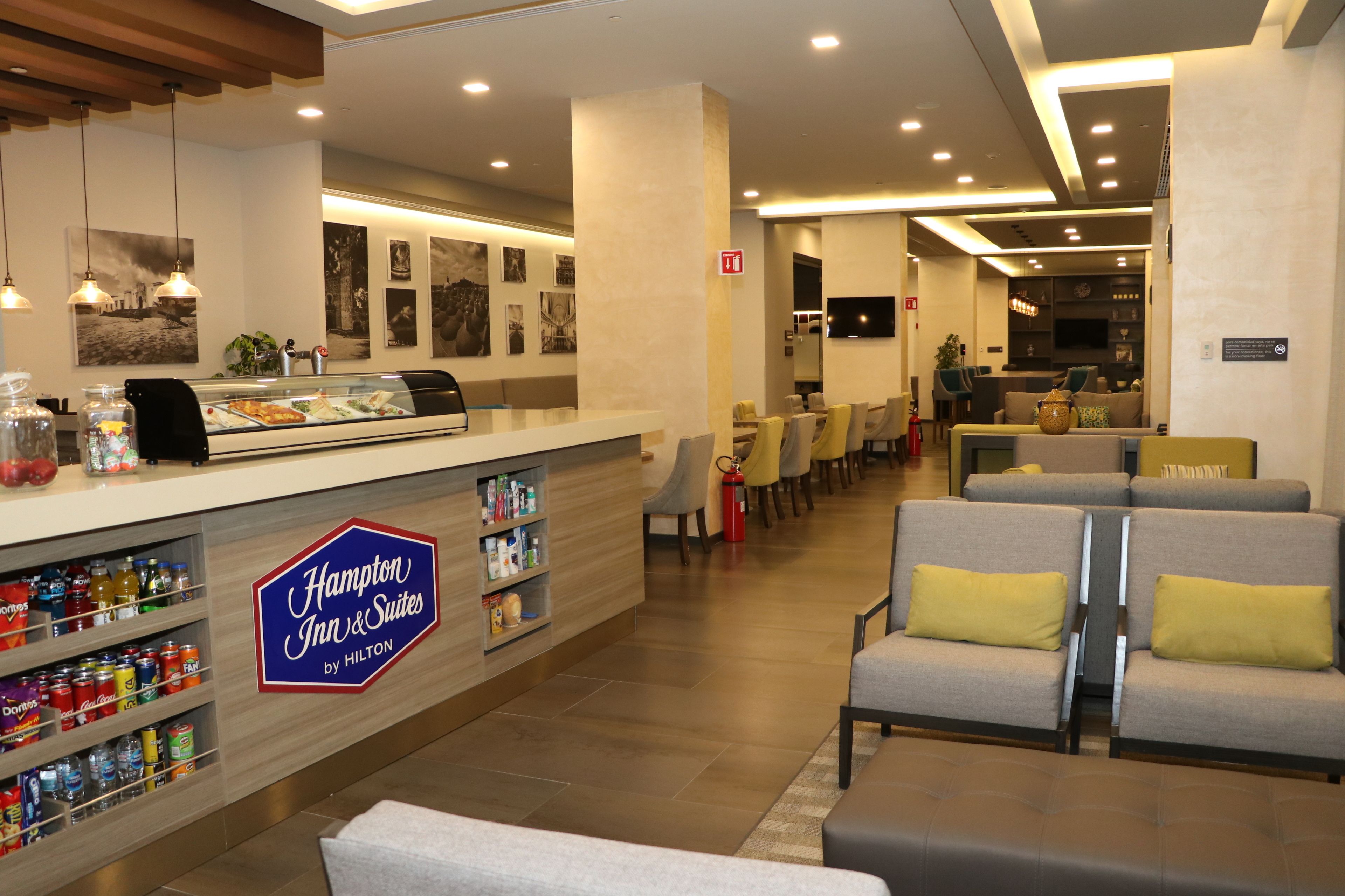 A Hampton Inn & Suites by Hilton lobby with a self-service breakfast bar, seating area, and a television screen.