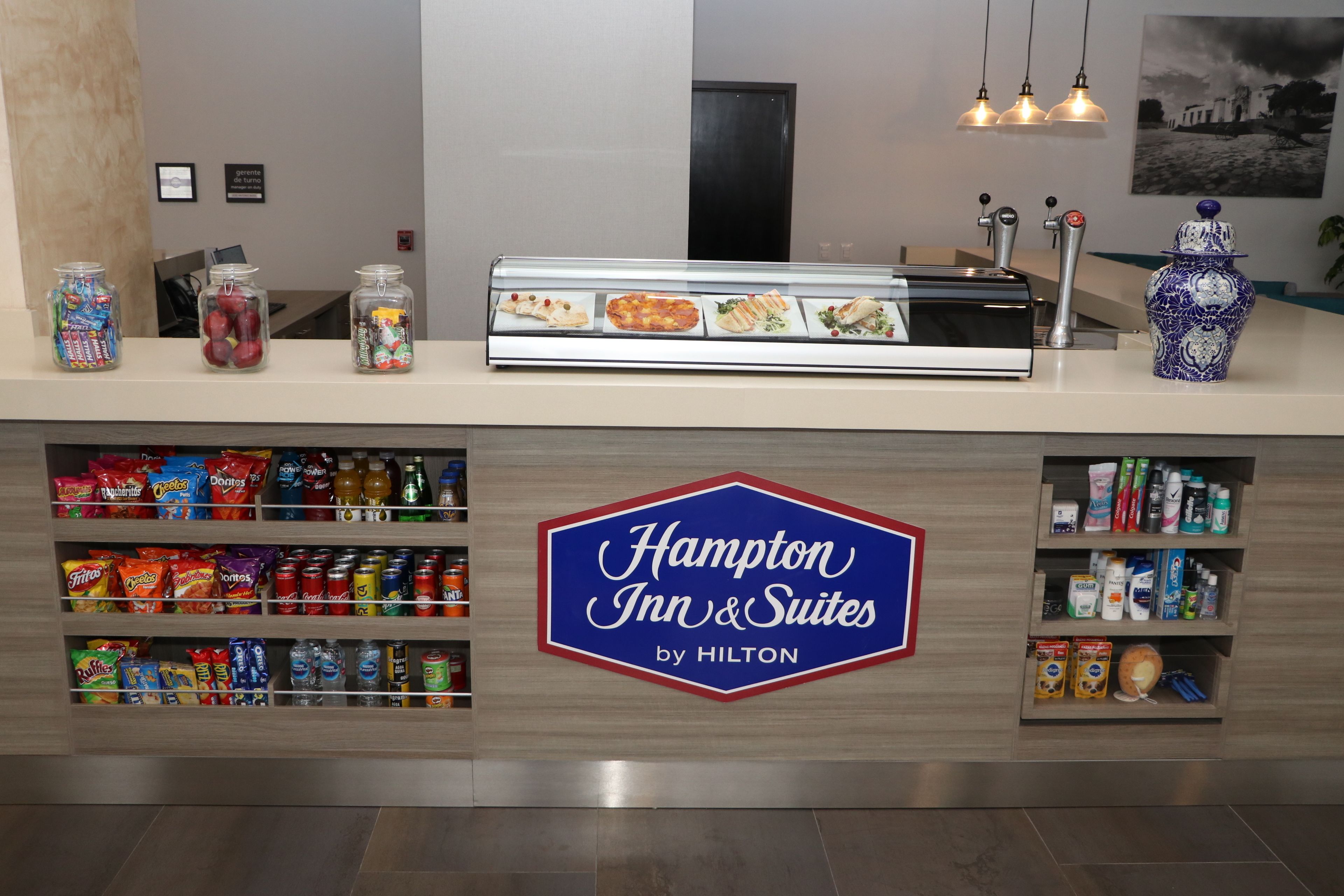 Zona de recepción de hotel con exhibición de bocadillos y letrero de Hampton Inn & Suites by Hilton.