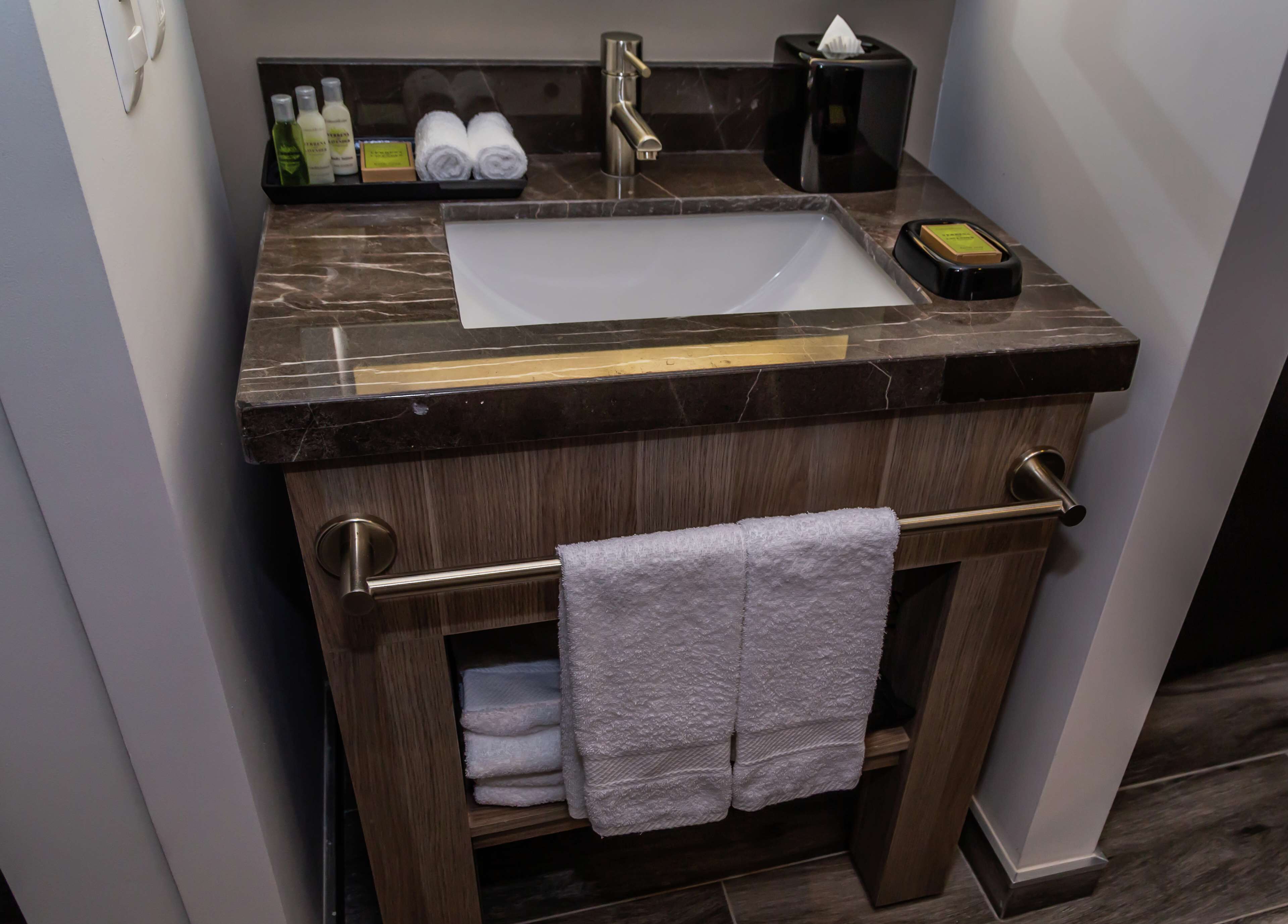 Un baño con encimera de mármol, un lavabo, toallero y estante con artículos de aseo.