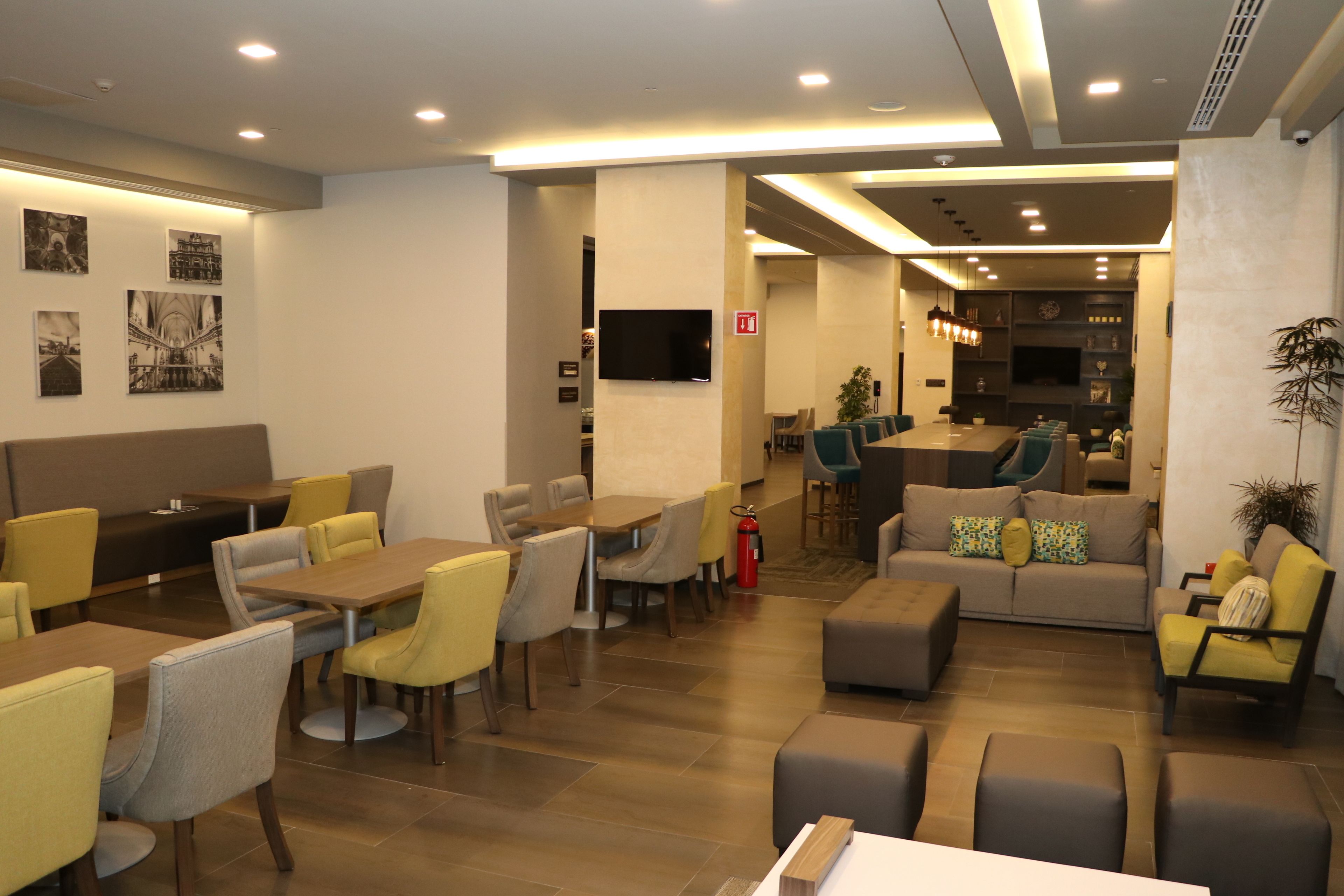 Un vestíbulo de hotel moderno con zona de comedor, sofás y un televisor montado en la pared.