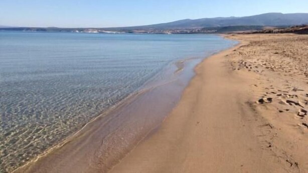 Ein Strand mit klarem Wasser, sandigem Ufer und in der Ferne Hügeln.