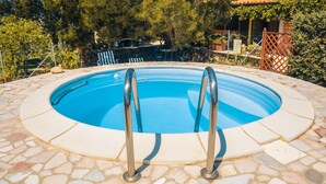 Ein runder Swimmingpool mit Metallleitern, umgeben von Steinplatten und Grünpflanzen.