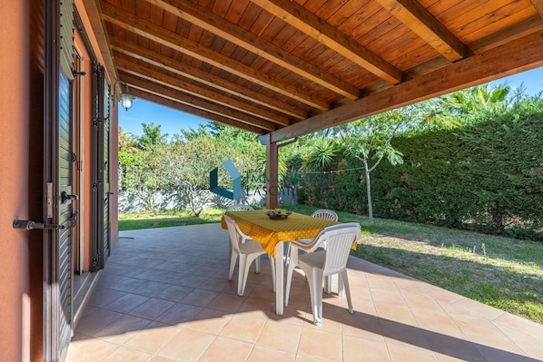 An outdoor patio with a wooden ceiling, a table with chairs, and a view of a garden with various plants.