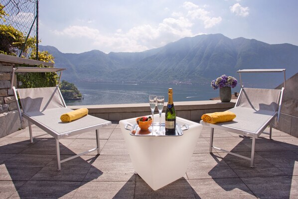 A terrace with two white lounge chairs, a table with a bottle and glasses, and a bowl of fruit.