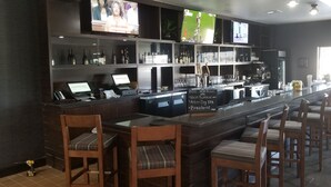 Zona de bar con una barra larga, varios televisores, estantes con botellas y una pizarra de menú.
