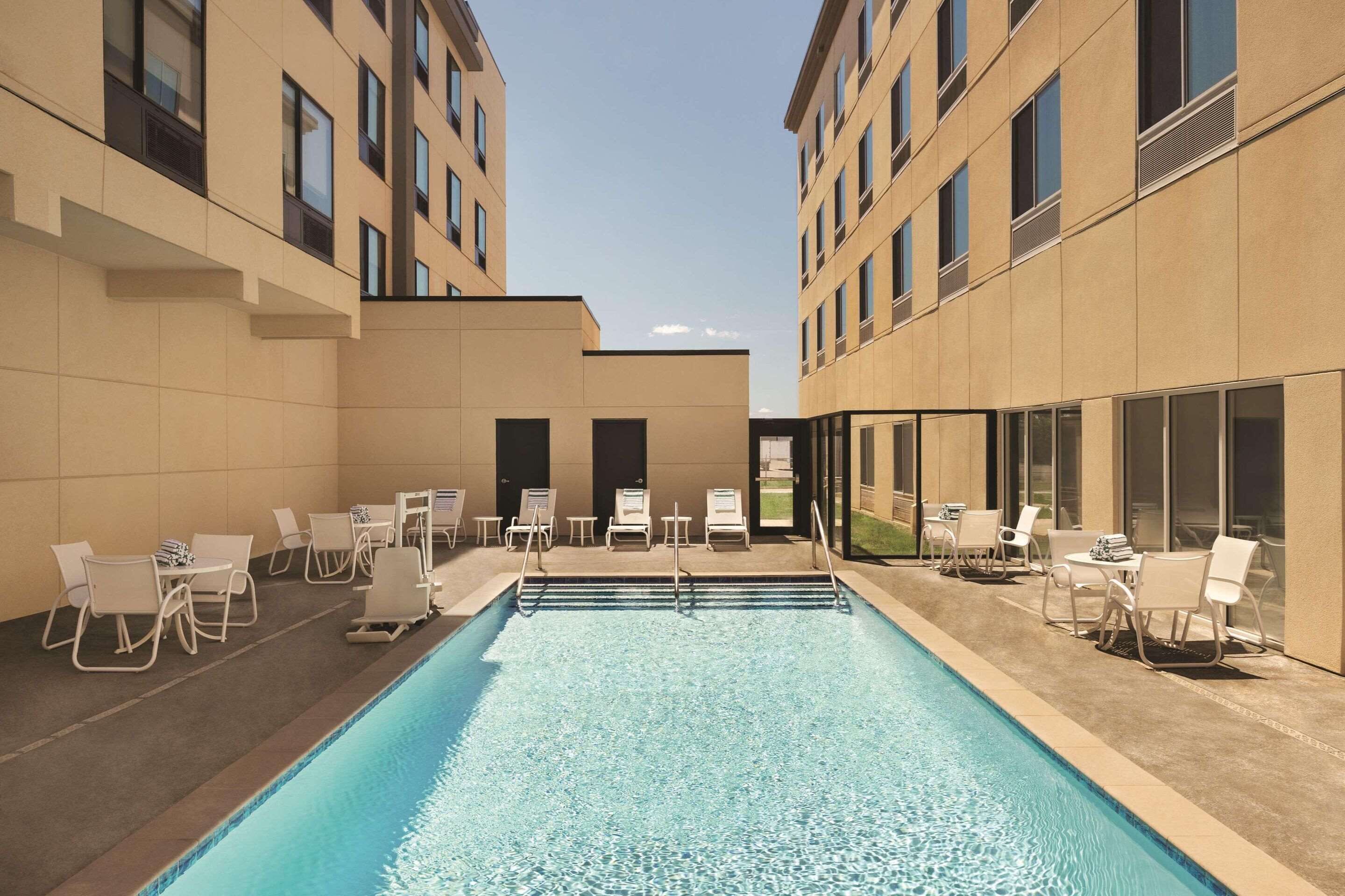 A swimming pool with lounge chairs and tables, surrounded by beige buildings with large windows.