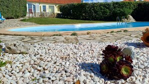 A swimming pool surrounded by pebbles and a garden area with a hedge and a building with a red roof. - Atlantic view house/front of the sea, private pool, Azenhas do Mar beach by walk (Colares)
