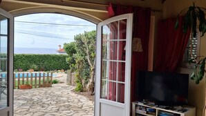 A room with a stone floor, a sofa, a coffee table, a TV, and a sliding glass door leading to an outdoor area with a pool and greenery. - Atlantic view house/front of the sea, private pool, Azenhas do Mar beach by walk (Colares)