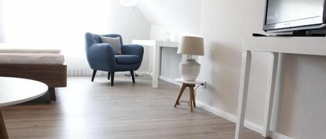 Una habitación moderna y luminosa con un sillón azul, una pequeña mesa redonda, una lámpara blanca y un televisor sobre un mueble blanco.