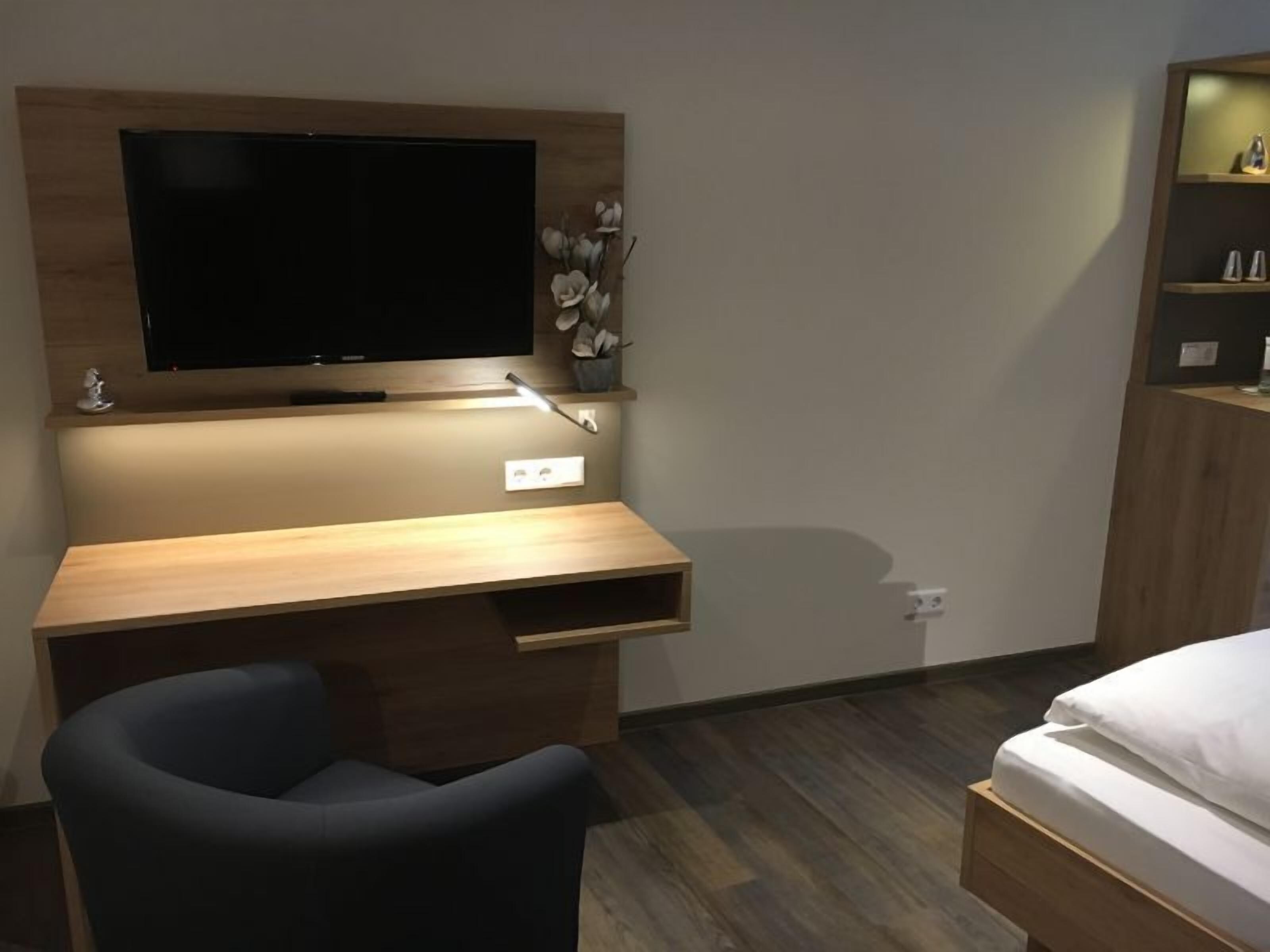 Une chambre d’hôtel moderne avec un bureau en bois, un téléviseur à écran plat, une chaise noire, un lit avec une literie blanche et une petite étagère ornée d’objets décoratifs.