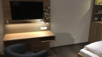 Une chambre d’hôtel moderne avec un bureau en bois, un téléviseur à écran plat, une chaise noire, un lit avec une literie blanche et une petite étagère ornée d’objets décoratifs.
