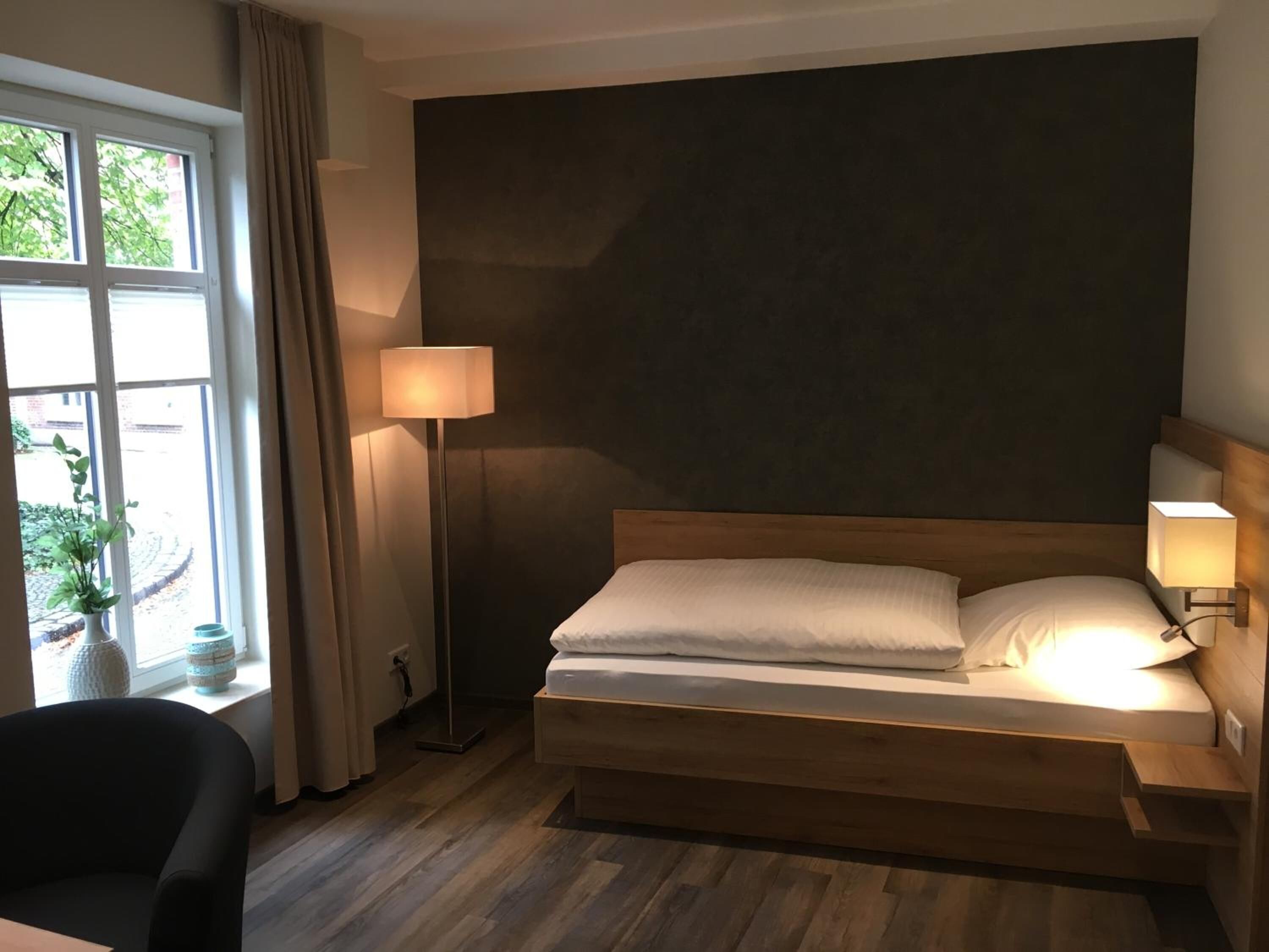 A modern hotel room with a wooden bed, a floor lamp, and a view of greenery through the window.