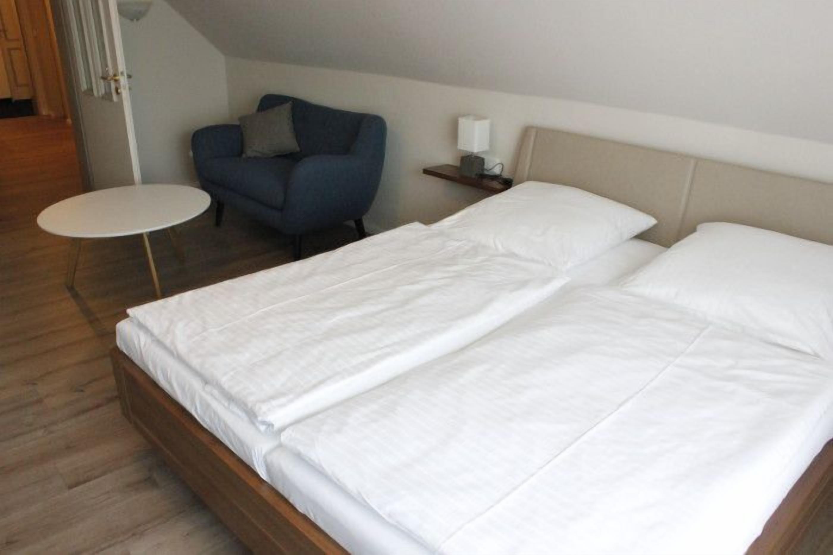 A bedroom with a double bed, a small round table, a blue armchair, and a bedside lamp.