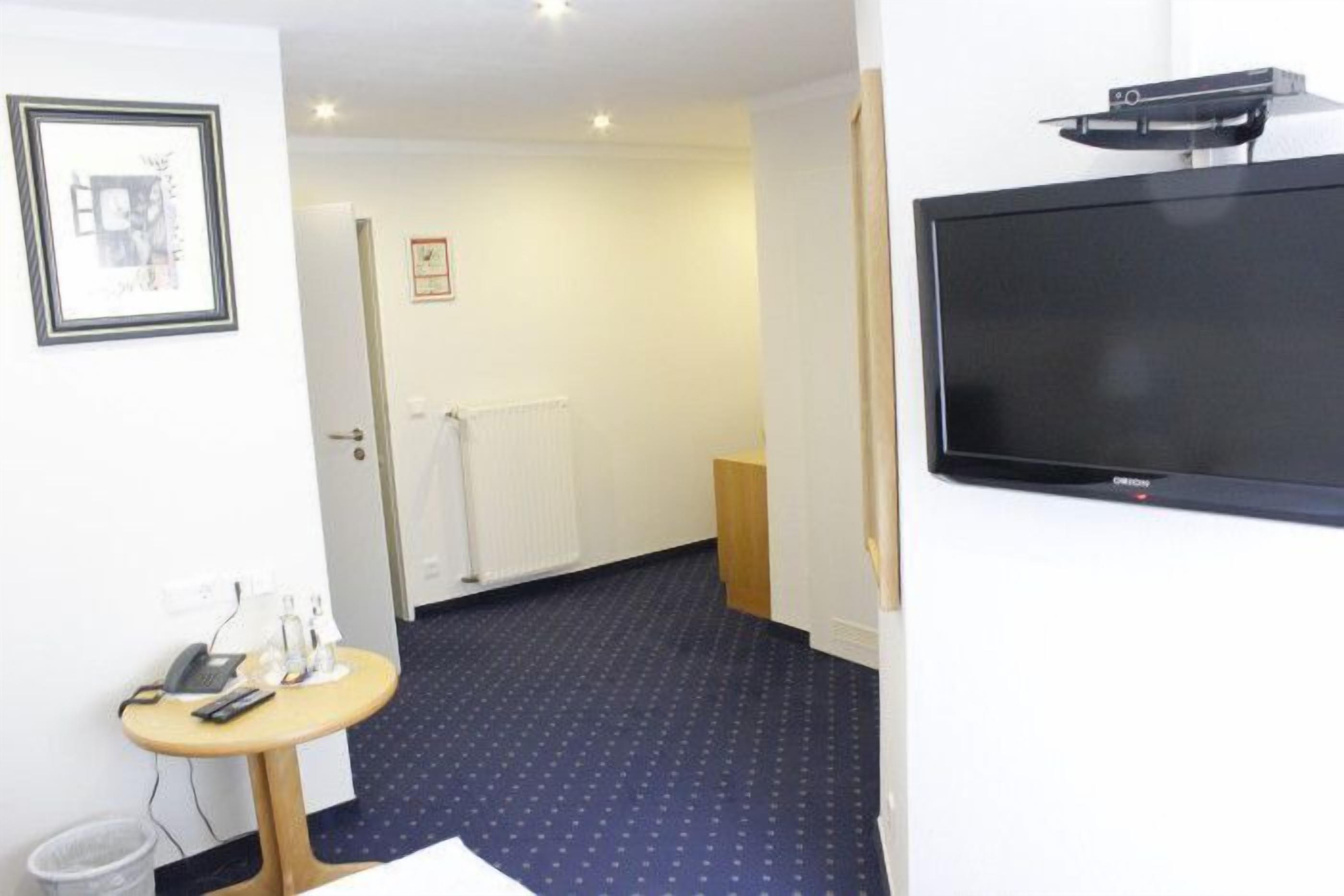 Ett hotellrum med en stor, platt-TV som är monterad på väggen, ett litet runt bord med en telefon och en lampa samt en mönstrad matta.