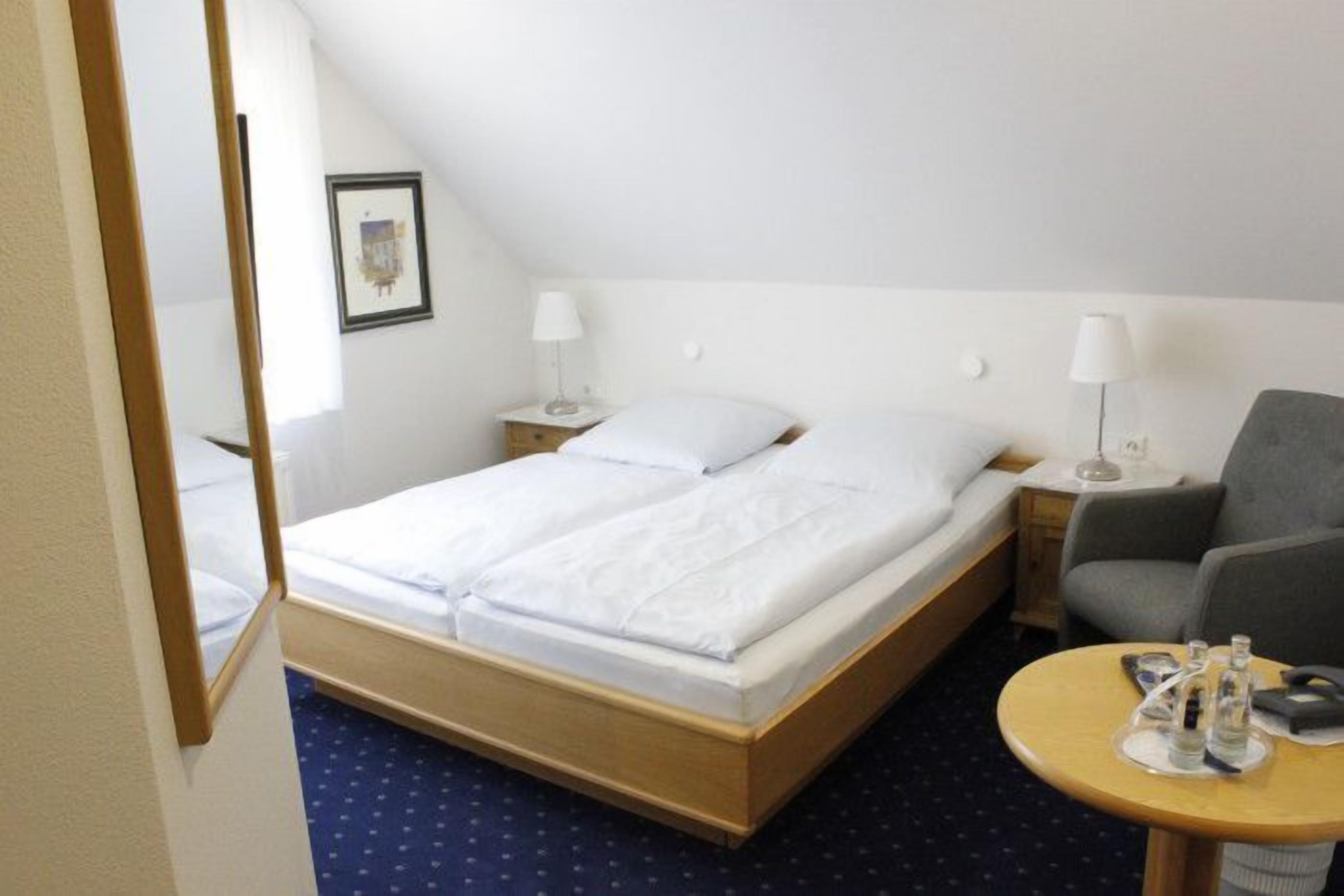 Habitación de hotel con una cama, dos mesitas de noche con lámparas, una silla y una pequeña mesa con una botella y un vaso.
