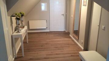 Une pièce avec un plafond en pente, un plancher en bois, une porte blanche, un radiateur, une petite fenêtre et un miroir accroché au mur.