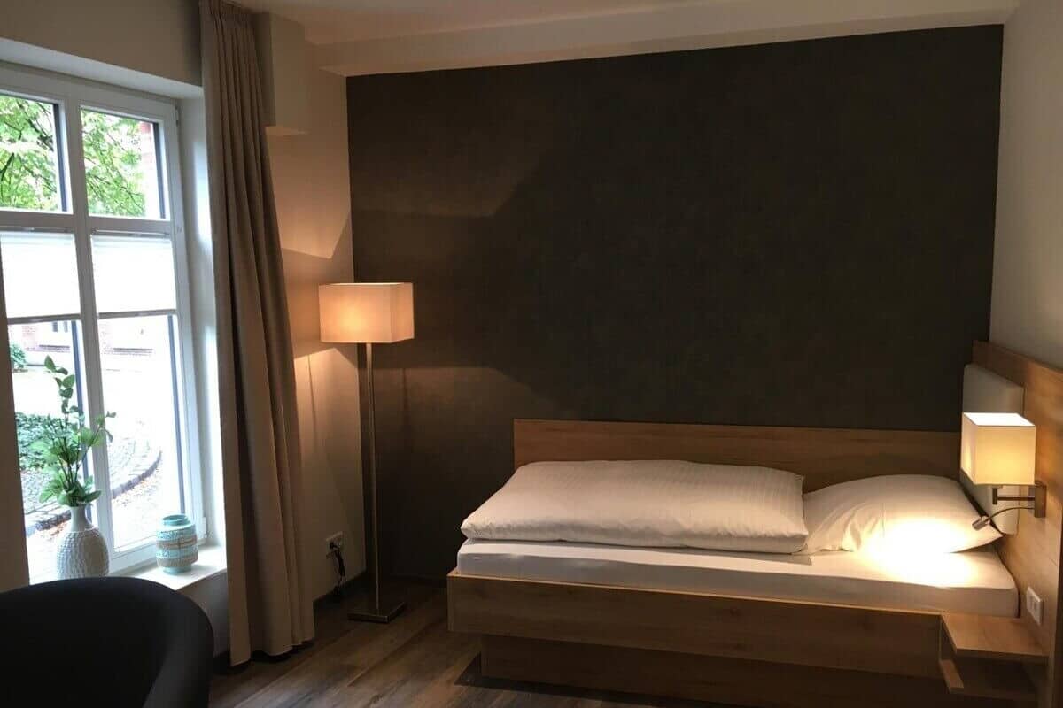 A hotel room with a bed, bedside lamps, a window with curtains, and a plant on the windowsill.