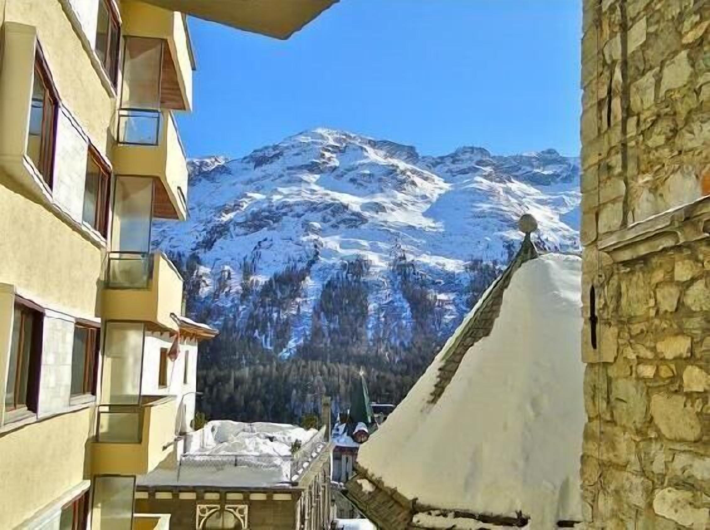 Vista de la montaña con picos nevados, un edificio de piedra y un edificio amarillo con balcones.
