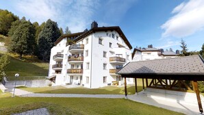 Un edificio a più piani con balconi, una zona esterna coperta e uno sfondo collinare.