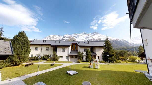 Un edificio a due piani con un ingresso centrale, circondato da un prato curato e una piccola area giochi. Sullo sfondo, una catena montuosa con cime innevate.