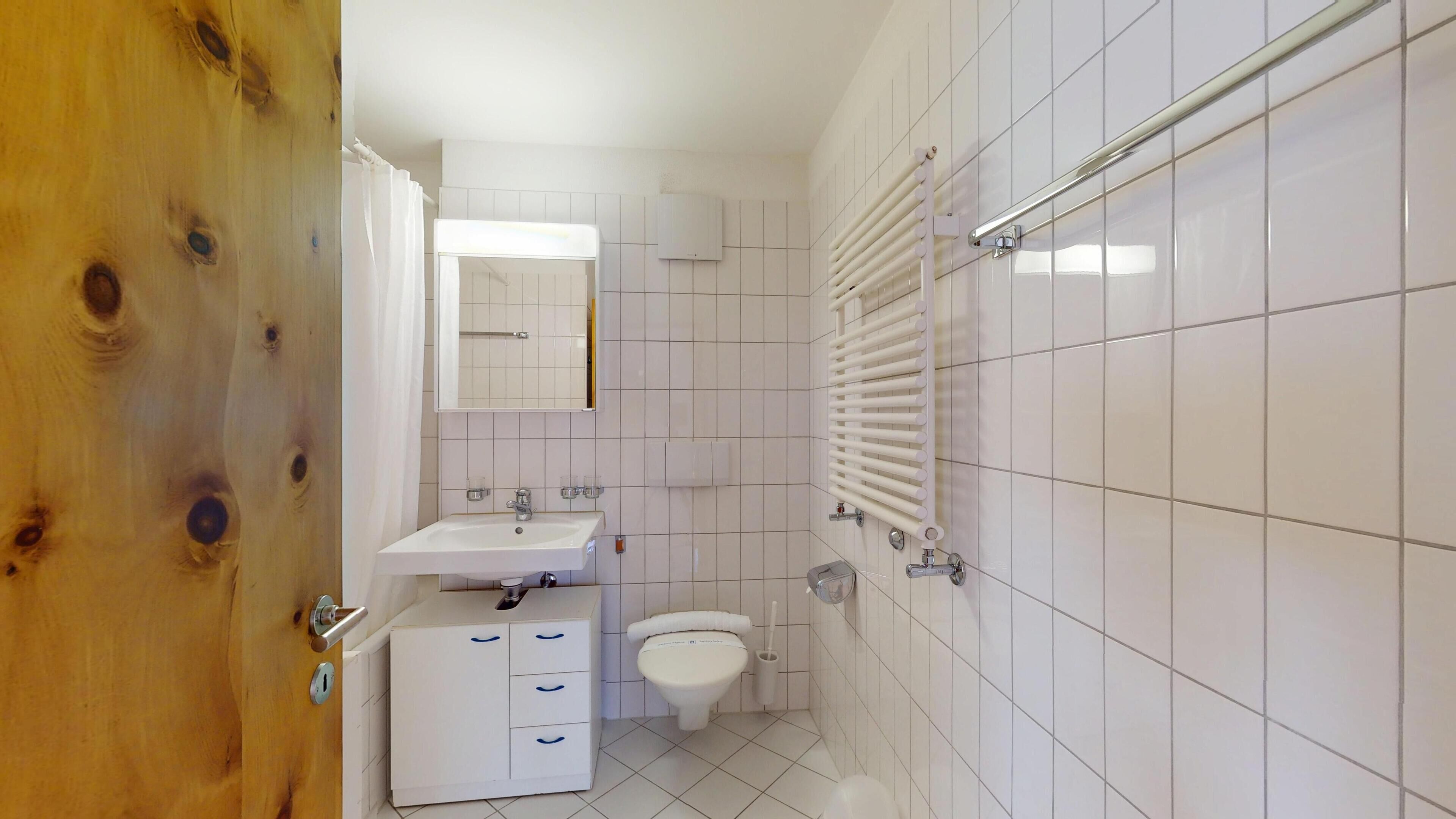 Un baño pequeño y limpio con azulejos blancos, un lavamanos, un inodoro y una zona de ducha.