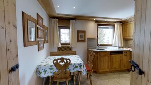 Uma cozinha acolhedora com armários de madeira, uma mesa de jantar com toalha de mesa florida e uma janela com cortinas.