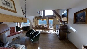 Ein geräumiger Wohnbereich mit Holzboden, einem Esstisch mit Stühlen, einem Sofa und einem Sessel. An der Wand hängt ein Gemälde und ein Fenster bietet einen Blick ins Freie.