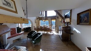 Uma sala de estar ampla com piso de madeira, uma mesa de jantar com cadeiras, um sofá e uma poltrona. Há um quadro na parede e uma janela com vista para o exterior.
