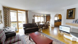 En stue med sofa, sofabord og fjernsyn. Der er et spiseområde med stole og bord. Værelset har store vinduer, og et køkken ses i baggrunden.