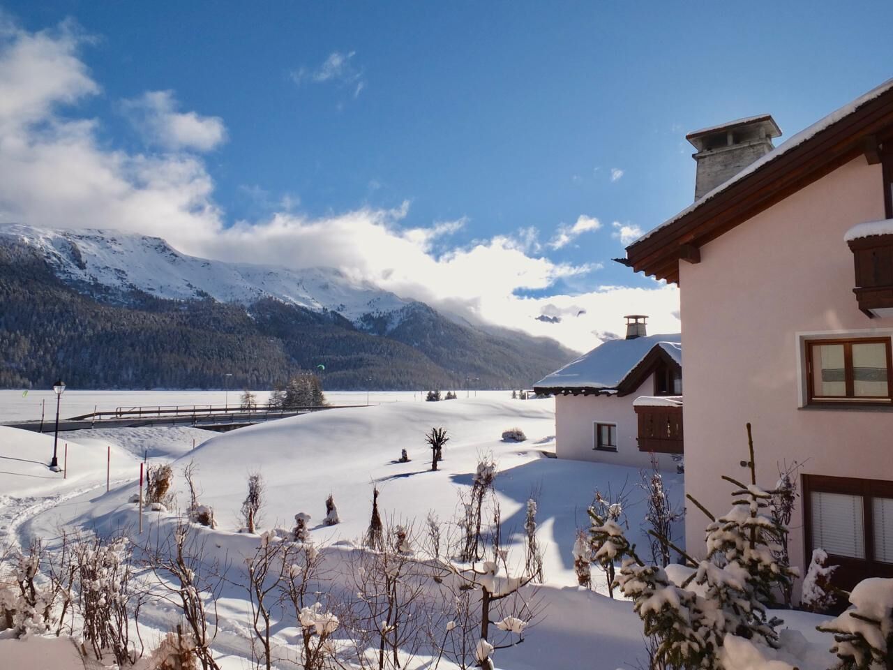 Paisagem coberta de neve com uma montanha ao fundo, céu claro e um edifício com telhado inclinado.