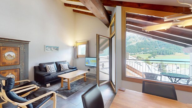 Una sala de estar moderna con un sofá de cuero negro, una mesa de centro de madera y un televisor que muestra 'LOG'. Un amplio ventanal ofrece vistas a un paisaje montañoso.
