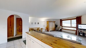 A spacious living area with a wooden dining table, a sofa, a rocking chair with teddy bears, a cabinet, and a television. - Spacious 3½-room apartment on the second floor, ca. (Silvaplana)