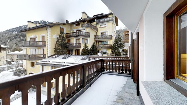 A balcony with a wooden railing overlooking a row of multi-story buildings with balconies, set against a mountainous backdrop. - Spacious 3½-room apartment on the second floor, ca. (Silvaplana)