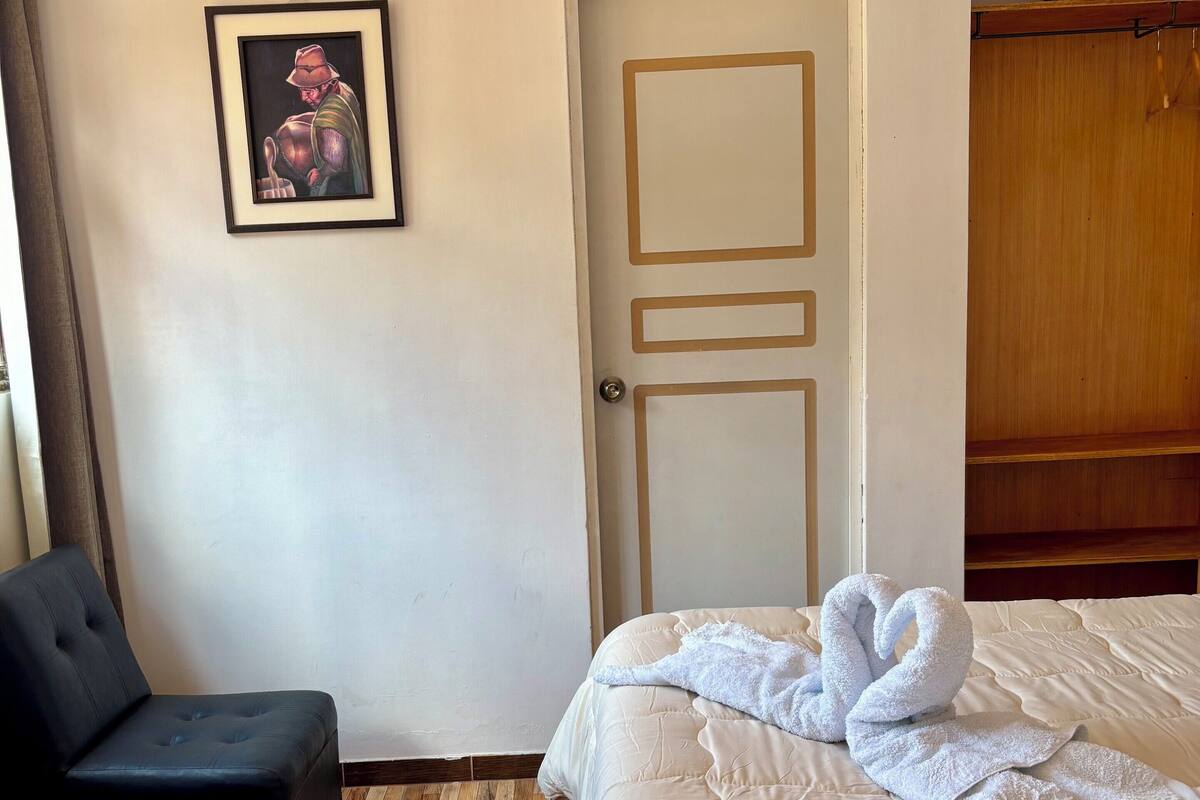 A hotel room with a bed, a black armchair, a small wooden table, a framed picture on the wall, and a wardrobe.