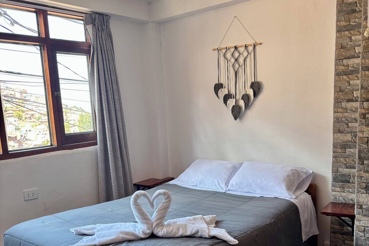 A neatly made bed with a heart-shaped towel arrangement, a macrame wall hanging, and a view of buildings through a window with curtains.