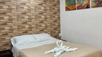 Quarto de hotel com cabeceira de madeira, cama com colcha bege e toalha em formato de coração sobre a cama. Há três quadros na parede.