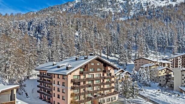 A multi-story hotel with balconies, surrounded by snow-covered trees and a mountainous backdrop.