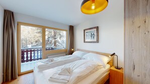A bedroom with a large window, a bed with white linens, a bedside table with a lamp, and a framed picture on the wall.