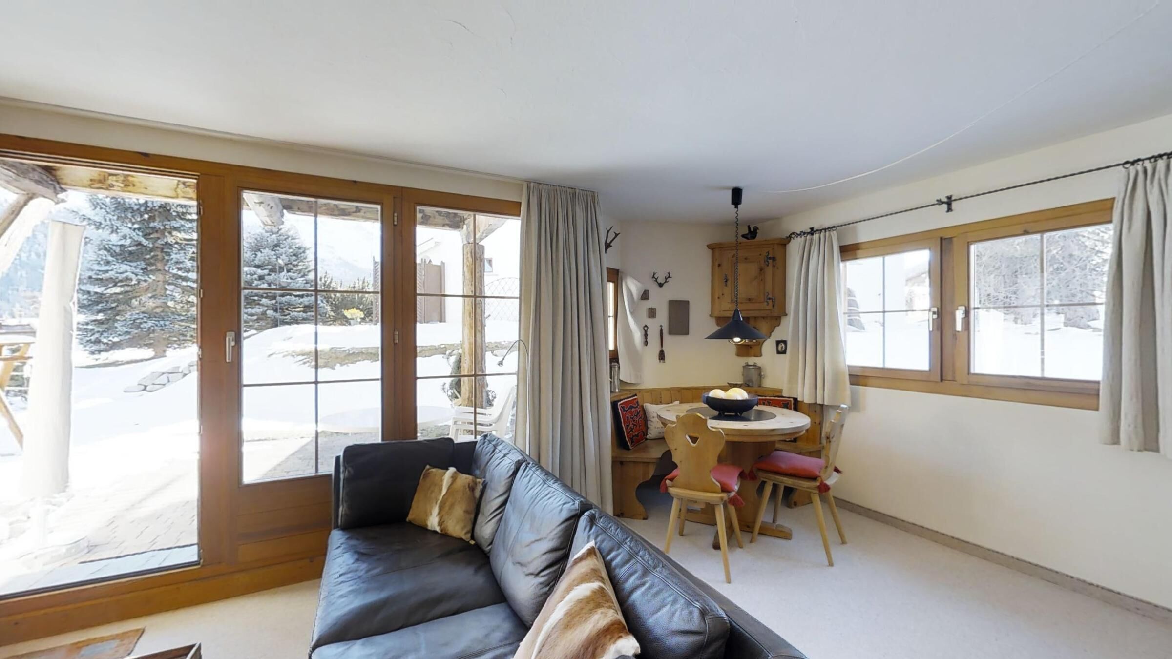 A cozy living room with a leather sofa, a dining table with chairs, and large windows offering a view of a snowy landscape.