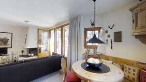 A cozy living room with a fireplace, a dining table with a bowl of fruit, and a large window with curtains. - 2½-room apartment on the ground floor, ca. (Silvaplana)