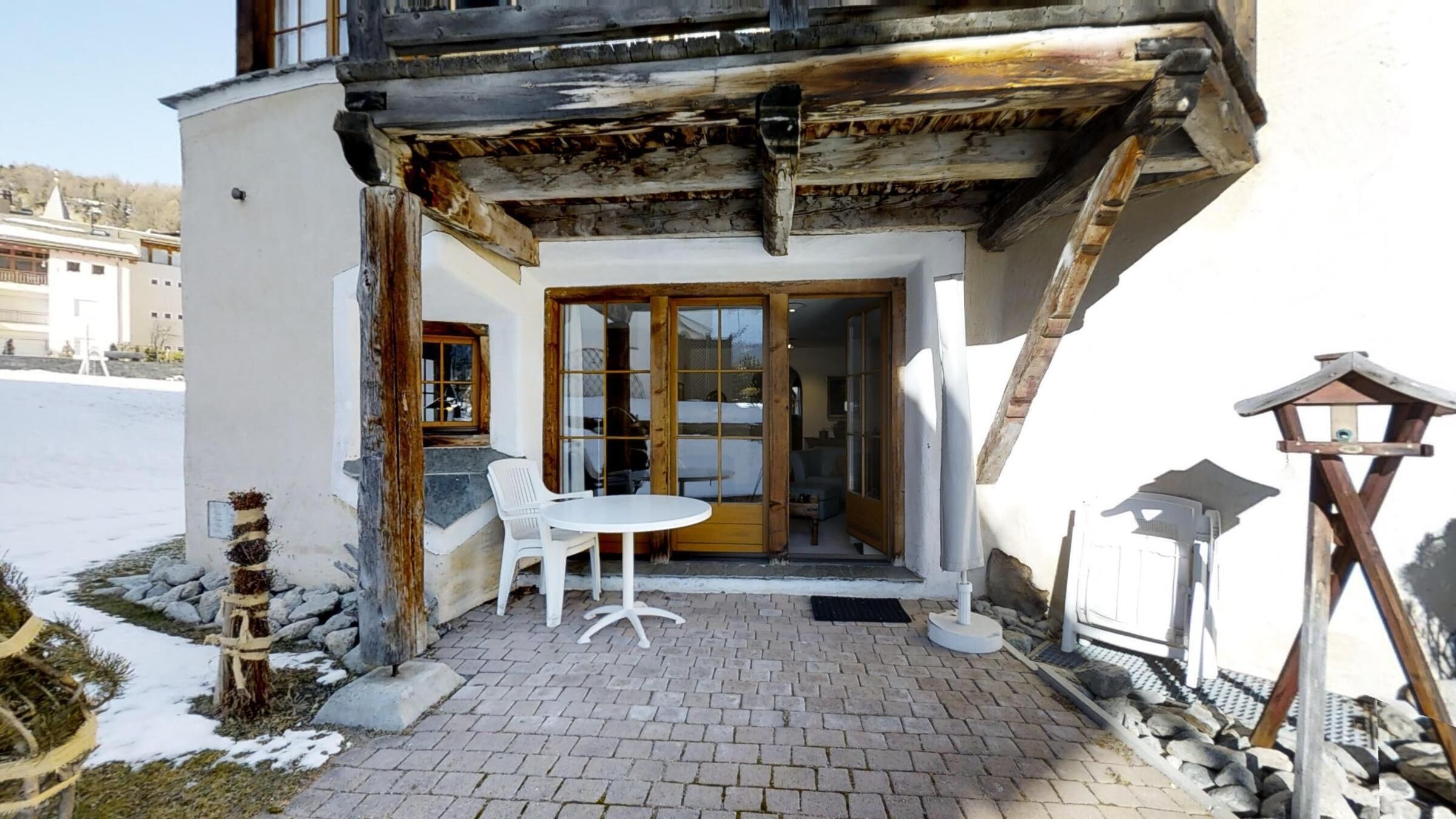 Un edificio con balcón de madera, una mesa y sillas blancas, y un camino de piedra.