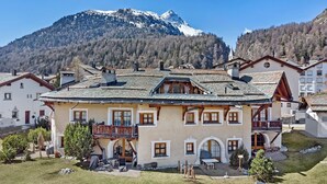 A traditional chalet with a sloped roof, wooden balconies, and a view of a snow-capped mountain. - 2½-room apartment on the ground floor, ca. (Silvaplana)