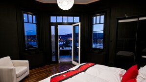 Ein Schlafzimmer mit einem großen Bett, einem weißen Sessel und Blick auf eine Stadtkulisse durch eine Glastür.