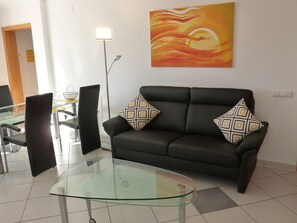 Una sala de estar moderna con una mesa de centro de vidrio, un sofá negro con cojines estampados y un cuadro abstracto grande en la pared.