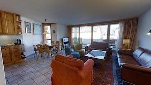 Una sala de estar con sofá, una mesa de centro y televisor. Hay un comedor con mesa y sillas. La habitación tiene ventanas grandes y vistas al exterior.