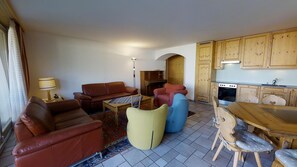 A living room with a brown sofa, a yellow ottoman, and a blue chair. There is a small table with a lamp, a rug, and a kitchen area with wooden cabinets and a stove.