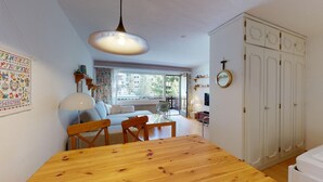 Habitación acogedora con una mesa de comedor de madera, un sofá y una ventana con vistas a edificios. Se observa un armario con espejo y una lámpara colgante.