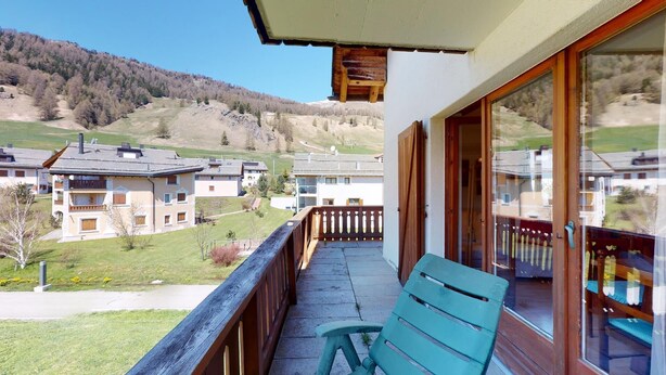 A balcony with a wooden railing, a green chair, and a view of a residential area with houses and a forested hill in the background. - 2-room apartment ca. (Celerina/Schlarigna)