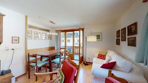 A cozy living room with a wooden dining table, chairs, a white sofa with red cushions, and a balcony with a view. - 2-room apartment ca. (Celerina/Schlarigna)