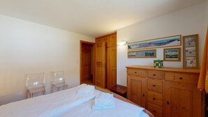 A bedroom with a bed, two chairs, a wooden wardrobe, and a hallway leading to a door. - 2-room apartment ca. (Celerina/Schlarigna)